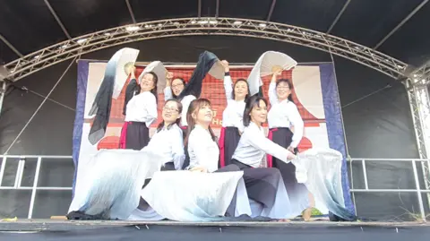 Wai Yee Hong Seven female Chinese dancers dressed in white shirts and long black skirts. They are waving white fans which are covered in scarves and they are all performing on a stage.