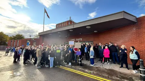 Cannock Chase Theatre Trust A crowd of protesters stand outside a red-brick building holding up several placards.
