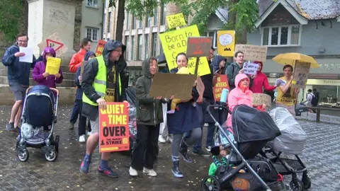 Women and men are striking for better paternity leave. They hold placards with slogans such as "Quiet time is over". It is a rainy day. Some have prams with them.