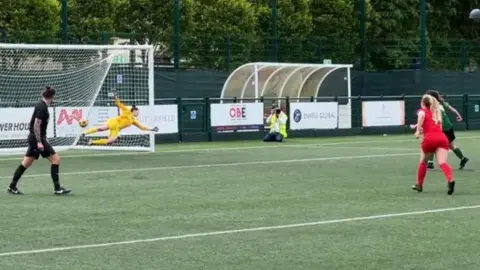 BBC A group of women, one team in red, the other in green, play football. The goalkeeper in yellow dives to their left to save the ball that is out of play.