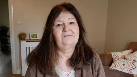 BBC Andrea, who has brown long hair and wears a brown cardigan, sitting in her living room with a neutral expression