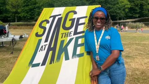 Bobsie Robinson is wearing a blue T-shirt and jeans and headband. She is standing in front of a large green and white striped deck chair in a field.