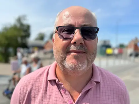 A head and shoulders photograph of Skegness Town Council member Paul Collins. He is wearing sunglasses and a pink shirt. He is mostly bald and has grey stubble.