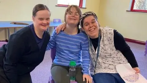 Three women sitting down hugging each other with bright smiles in a meeting room. 