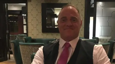 Supplied Ray McDonald is sitting in a bar. He is a middle-aged man wearing a white shirt, a dark pink tie and a dark grey or black waistcoat. He has a receding hairline and is smiling. Behind him are green chairs grouped around tables with a large mirror on the wall.