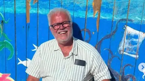 Tony Francis/Instagram Mr Francis smiles for the camera while sitting next to a beach hut. He has grey hair and a beard and is wearing a green and white striped shirt