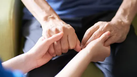 A close up of two people holding hands, one pair of hands visibly older than the other. The two people appear to be sitting down and wearing dark colours.