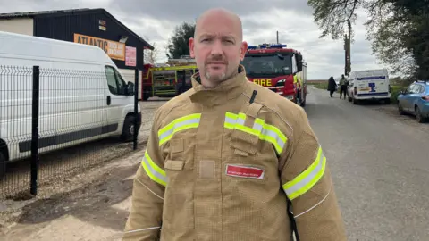Owen Sennitt/BBC Ryon Martin wears a yellow fire fighter jacket with hi-vis stripes. He is standing in front of the industrial building and a fire engine in a rural location.