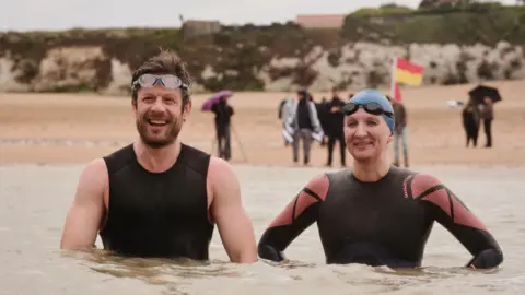 Actor James Norton and swimmer Rebecca Adlington in the sea, with a beach behind them. Both are wearing wetsuits and have goggles on their heads.