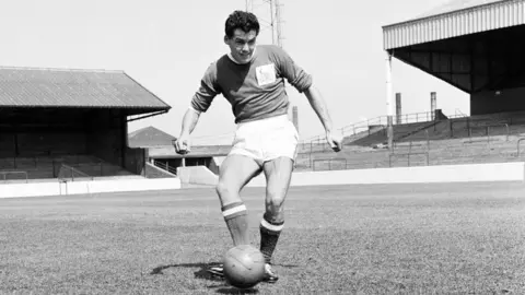 Getty Images Frank Wignall is pictured in his Nottingham Forest kit with a ball at his feet. The photograph is taken in the middle of the pitch. The football ground is deserted his body angled with one leg bent to kick the ball. His expression is one of focus and concentration as he looks ahead across the pitch 