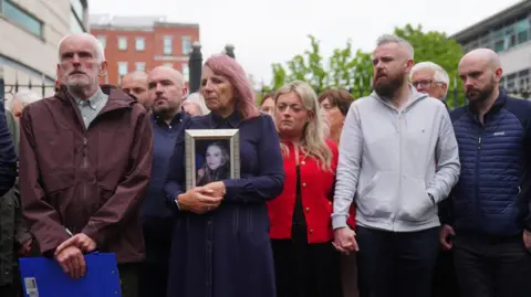 PA Media A group of solemn people stand together outdoors, with a woman in the centre holding a framed photo of a young woman while others hold hands beside her.