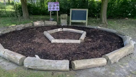St Augustine's Academy Wooden logs arranged in a circle around bark chippings, next to some trees and an outdoor information board about Forest School. 