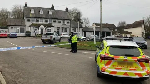 Dan MacLaren/BBC Police stand outside the pub, which is taped off. A police car is in the foreground.