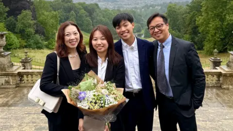 Cherry Lee A family of Hong Kong nationals are standing at Prior Park in Bath wearing formal clothing. One is holding a big bunch of flowers. Behind them is some elaborate stonework and a tree-lined hill