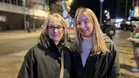 Two women, one wearing glasses, are outdoors and standing to pose fo rthe photo. They are both wearing black coats.
