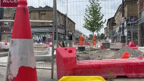 A picture of roadworks in Brighouse. There is a cone to the left and metal fencing, there are two workmen in hi-vis and helmets with rubble and gravel around them. There are shops in the background.