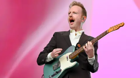 A man playing guitar is wearing a black suit and whit shirt. He has ginger hair and a moustache, and has his mouth open as he plays