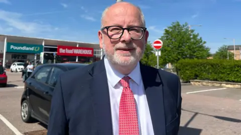 Steve Yemm, Labour MP for Mansfield, stood in a retail park car park with a red tie, white shirt and dark blue blazser