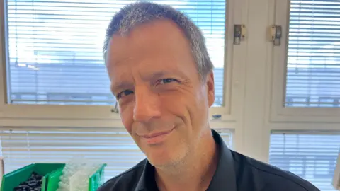 A man in a lab smiling at the camera. He has short grey hair and is sat in front of windows covered with horizontal blinds.