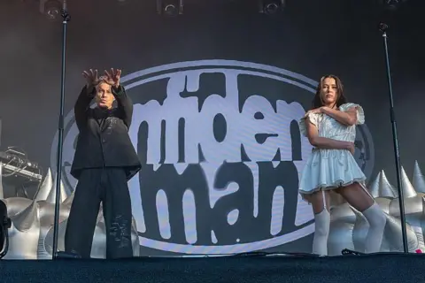 Getty Images A man in a baggy black suit holds his arms towards the crowd. He is stood next to a woman in a white dress and white tights. The words confidence man are spelled out on a screen behind the pair