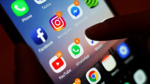 Close-up of a smartphone screen displaying social media and messaging apps, including Instagram, WhatsApp, Facebook, YouTube, Snapchat, and Messenger. Several apps show notification badges, and a finger is touching the screen.