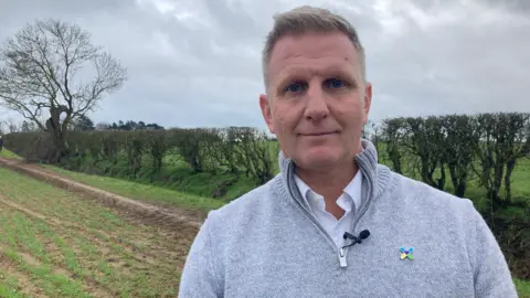 Chris Bailey from the Air Ambulance Service wearing a grey top and standing on land at Catthorpe in Leicestershire with a hedge line and trees in the background. 