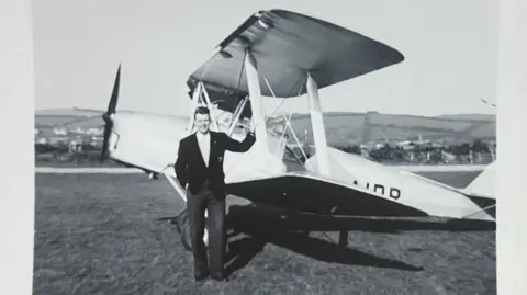 Handout A black and white photograph of a man standing in an airfield at an airplane.