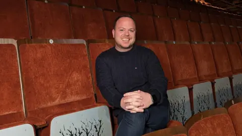 Aylesbury Waterside Theatre Grant Brisland, director, Aylesbury Waterside wearing a navy cable jumper and jeans sitting on a red, velvet theatre chair. He is smiling with short dark hair 
