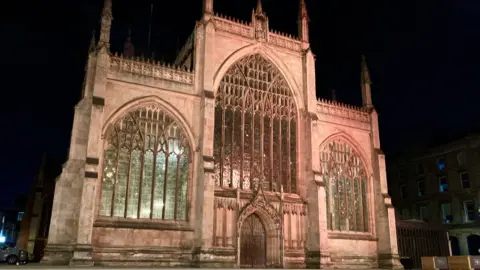 Hull Minster by night with three large glass windows and an entrance at the front. The Minster is lit up on the outside and inside.