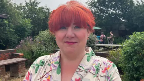 BBC News A woman wearing a floral top looks at the camera. She has red hair and a fringe and is standing in front of some benches and trees. 