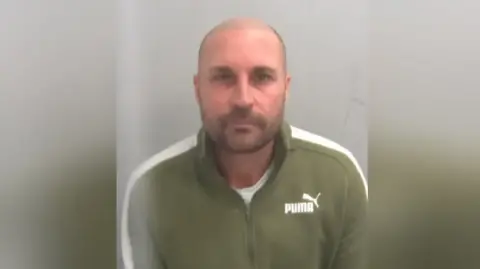 Essex Police A mugshot of David Richards. He is bald with stubble and is wearing a green sip-up jumper with a Puma logo to the right.