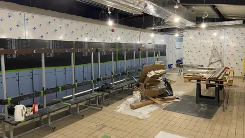 A changing rooms facility undergoes renovation. Blue lockers and seats and pegs can be seen on the left with a white kettle and PG tips box perched on the side. Cardboard boxes can be seen in the middle of the room along with a wooden panel on a table.