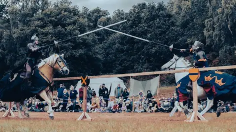 RJJonesPhotography Two jousters in full medieval knights armour and helmet are drawing sword on horseback as they move across a fence towards each other. A crowd can be seen sat on grass behind fencing.