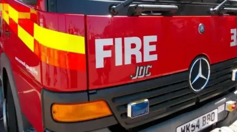 Close-up shot of a red fire engine from Cornwall Fire Service. The word fire is written on the front in large white capital letters. A large Mercedes Benz badge is also on the front of the vehicle.