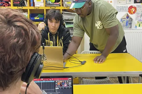 Oyindamola Odelabu A man leans over a laptop with a teenager sitting looking at the screen. In the foreground is the back of a person's head with headphones on.