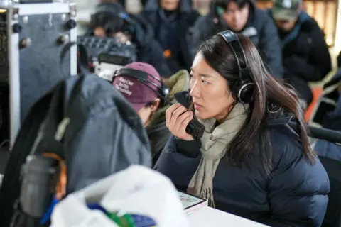 BBC/Hosu Lee A woman with dark hair wearing headphones speaks into a walkie-talkie.