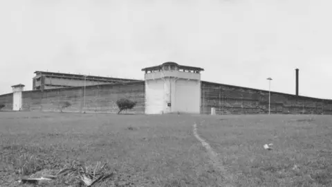 Getty Images A black and white image from the outside of a large prison. There are two watchtowers with high walls between them. A large building is behind the walls.