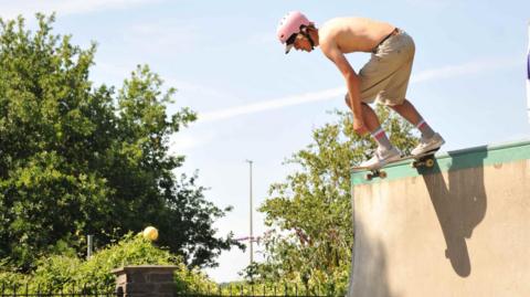 Man skateboarding without wearing a top, with pink helmet on. He is skating at park near trees.
