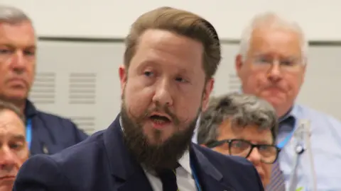 A man who is mid-talking during a meeting. He has light brown hair and a thick beard. His mustache has been turned upwards at the ends. He wears a blue suit, white shirt and dark blue tie. Other men sit behind him listening. He ha