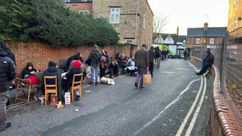 Bella Barnes A road with a fence on one side and a brick wall on the other, with a row of people lining one side. Some are sitting on dining chairs and most are wearing hats and coats, with food and bags at their feet.