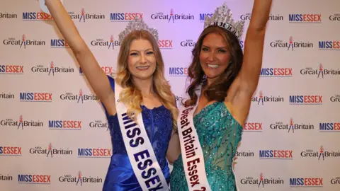 Cutter Creatives Claire Redgrave and Abby Squires standing in front of a white backdrop, with Miss Essex and Miss Great Britain logos on it. Claire is wearing a blue sequined dress, a tiara and a white sash, and has long blonde hair. Abby is wearing a teal sequined dress, a tiara and a white sash, and has long brunette hair.