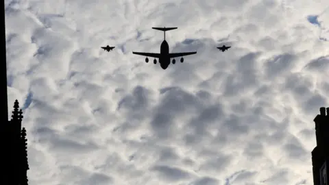 A large cargo plane in silhouette flanked by two of the Red Arrows against a cloudy sky