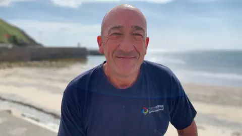 Ray is a bald man, wearing a dark blue t-shirt, smiling in front of Laxey beach behind him on a calm and sunny day.