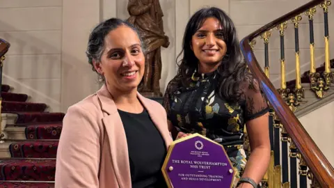 The Royal Wolverhampton NHS Trust Two women standing on a staircase holding an award. The one on the left smiles showing her teeth and wears a pink jacket over a black top. On the right, the other woman has long black hair past her shoulders and also smiles showing her teeth. She wears a black dress with a flower petal design. Between them, they hold the award - a purple octagonal piece with gold edging and writing on it including "Royal Wolverhampton NHS Trust".