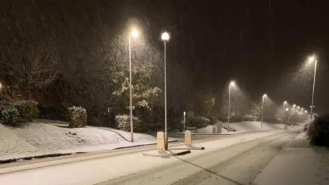 Snow on a road in Okehampton. Footprints are in the snow. It is dark. Lampposts light up the street.