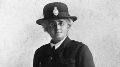 A black and white photo shows a woman looking directly at the camera. She has short hair and glasses. She's wearing a police officers hat, and is in uniform. 