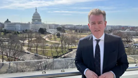 Mark Carruthers berdiri di balkon dengan Gedung Capitol AS di latar belakang. Mark mengenakan setelan angkatan laut di atas kemeja putih dan dasi navy.