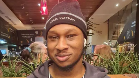 Khaleed Oladipo wearing a black hat and zip-up jacket, smiling. 