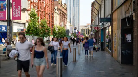 Pedestrians walk along a busy shopping street in Glasgow city centre, with retail stores, trees, construction scaffolding and modern high-rise buildings lining the pedestrianised thoroughfare.