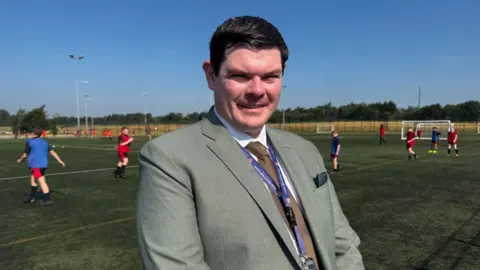 Jonny Humphries/BBC A middle aged man with black hair, and wearing a light grey suit, stands on the edge of an artificial pitch where schoolchildren are playing football
